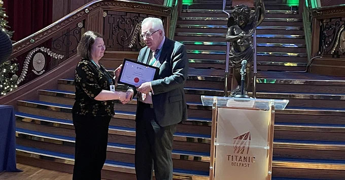 Two individuals presenting an award at an awards ceremony. One person is handing a certificate to the other on a stage beside a lectern with the Titanic Belfast logo, in front of an elegant staircase. 