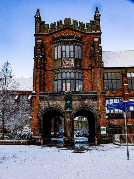 Snow-covered Newcastle University archway and building, with Gothic architectural features and a vibrant blue sign visible.