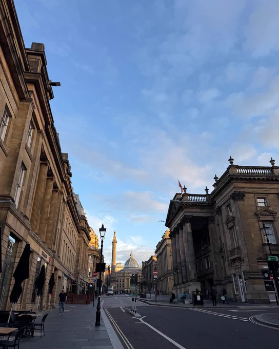 Early morning view of Grey Street in Newcastle, leading towards the iconic Grey's Monument and the dome of the Theatre Royal visible under a clear blue sky.