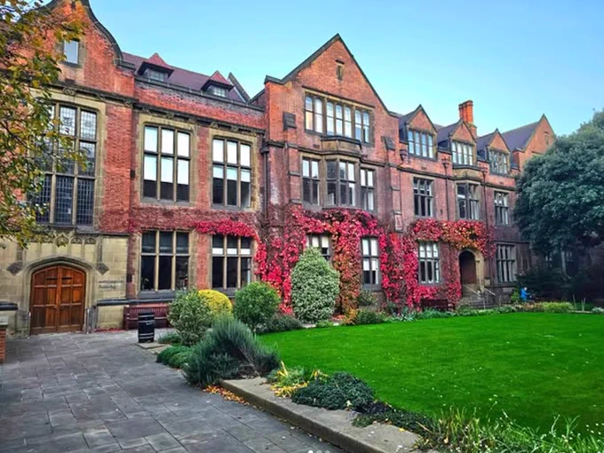 Exterior view of a historical brick building with large windows, adorned with ivy, partially covered in red and green leaves. The building is surrounded by a manicured lawn and small garden areas, under a clear sky.