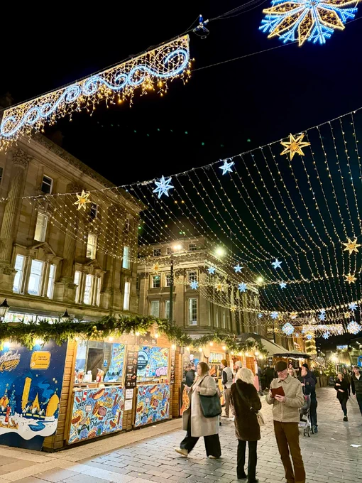 Festive decorations and lights hang above a bustling street market at night, with visitors walking by and stands featuring colorful displays.