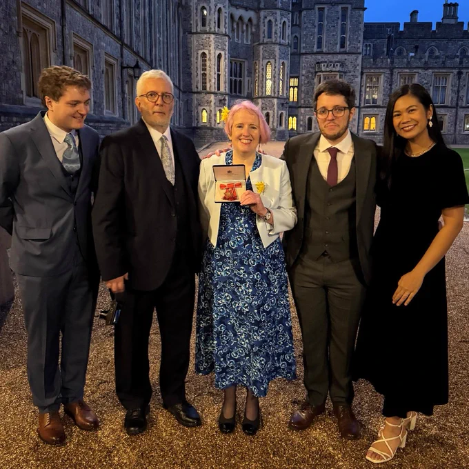 Five individuals posing with an award in front of Windsor Castle at night. One person is holding a red award box. They are dressed in formal evening wear.