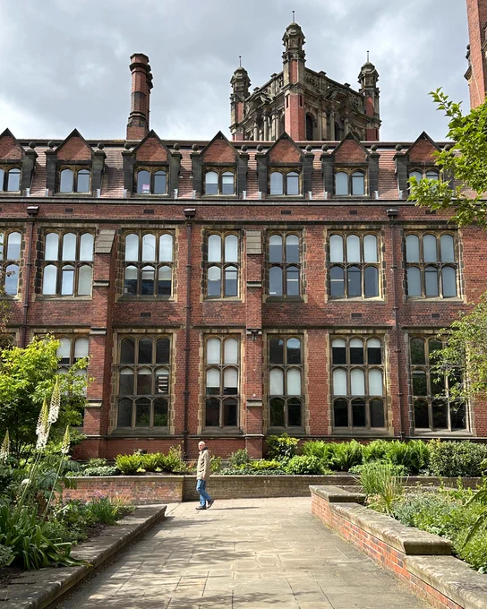 A person walking in front of the historic and ornate building at Newcastle University, surrounded by lush greenery.