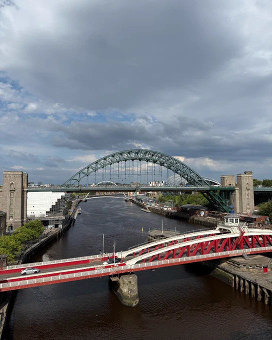 An elevated view of the Tyne Bridge over the River Tyne, flanked by modern structures, with the Swing Bridge in the foreground under a partly cloudy sky.