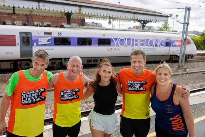 Five LNER colleagues in sportswear, some CALM branded, together on a station platform with the 'You Belong' Azuma train behind.