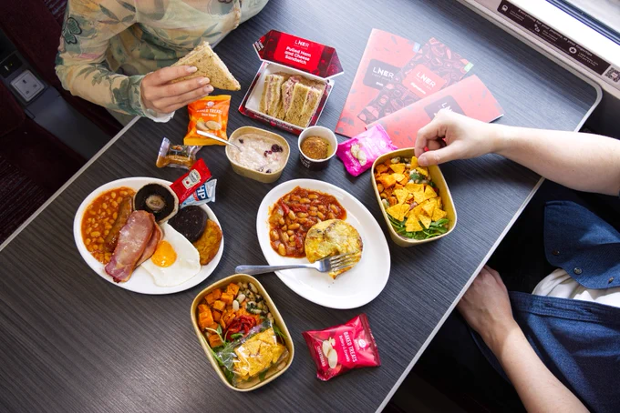 A table displaying a variety of meals from the new LNER First Class Menu, onboard the First Class carriage of an LNER Azuma service. There are 2 customers enjoying the food. 