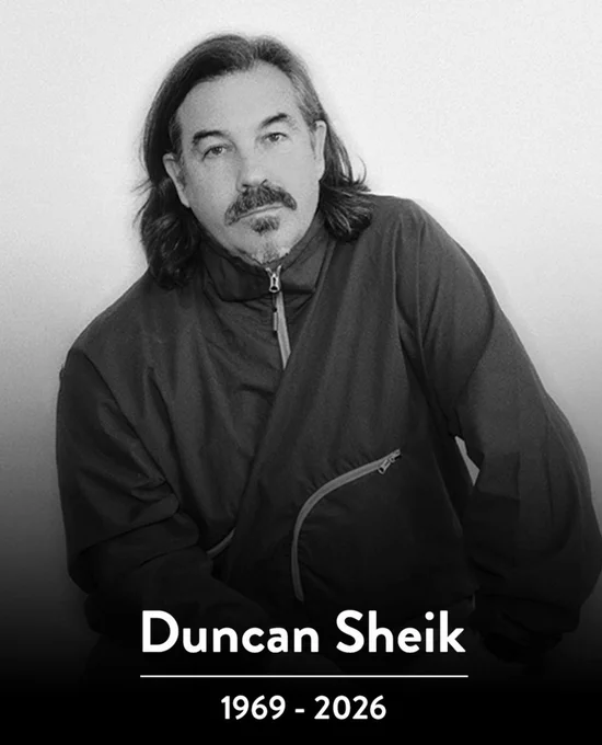  A black and white image of a man looking into the camera. He has long hair and a mustache and is wearing a jacket and tie. Below the image, white text reads "Duncan Sheik 1969 - 2026"